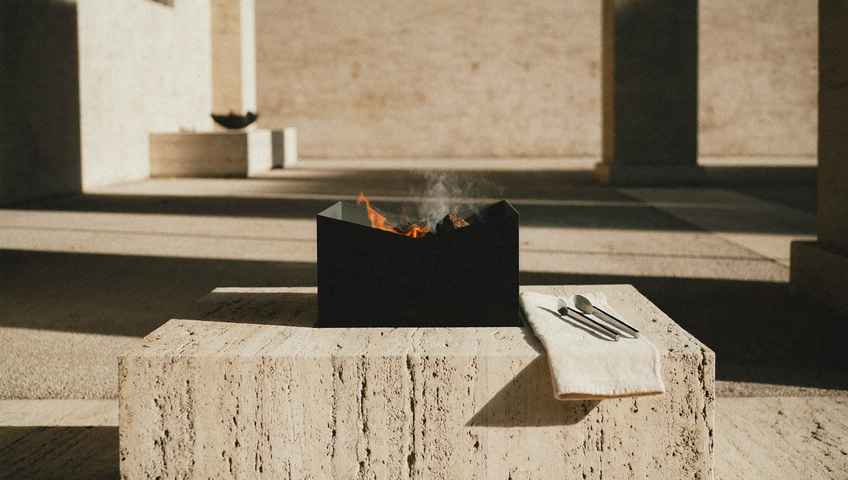 Griglia a carbone in acciaio nero opaco su un basso plinto di travertino, con braci accese all’interno e pinze in acciaio spazzolato appoggiate su lino piegato, nella luce radente del tardo pomeriggio.