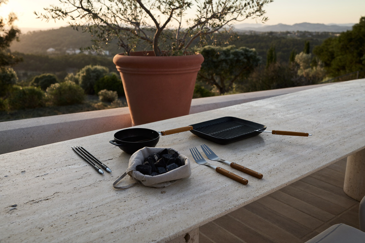 Lungo tavolo in travertino su una terrazza all’ora dorata con griglia in acciaio nero, padella in ghisa, spatola e pinze con manico in olivo, spiedi in acciaio e un sacchetto di lino con carbone di legna; colline sfocate sullo sfondo.